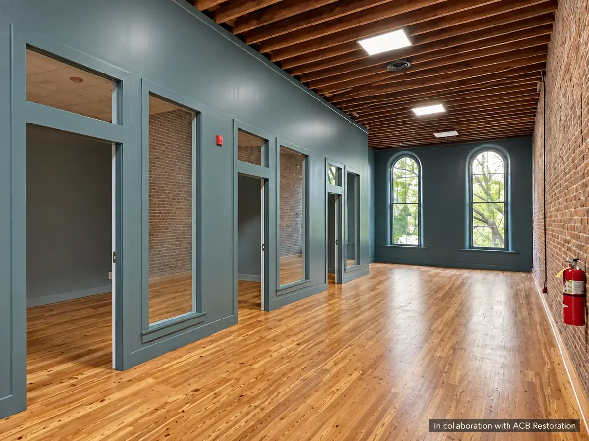 Office interior with exposed brick, timber ceiling joists, slate-blue partitions and tall arched windows. In collaboration with ACB Restoration.