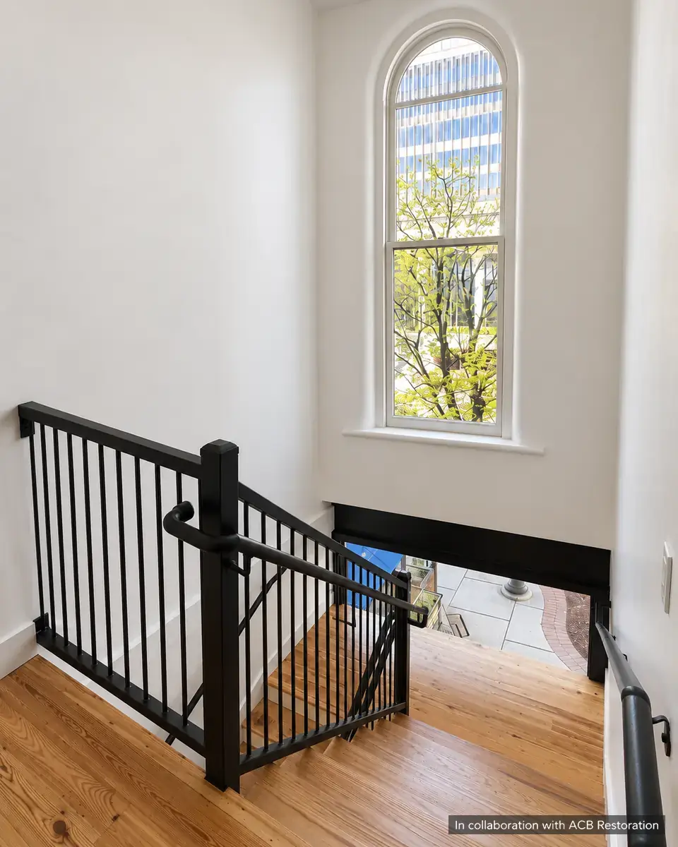 Tall arched window lighting a timber stair with a black metal railing. In collaboration with ACB Restoration.
