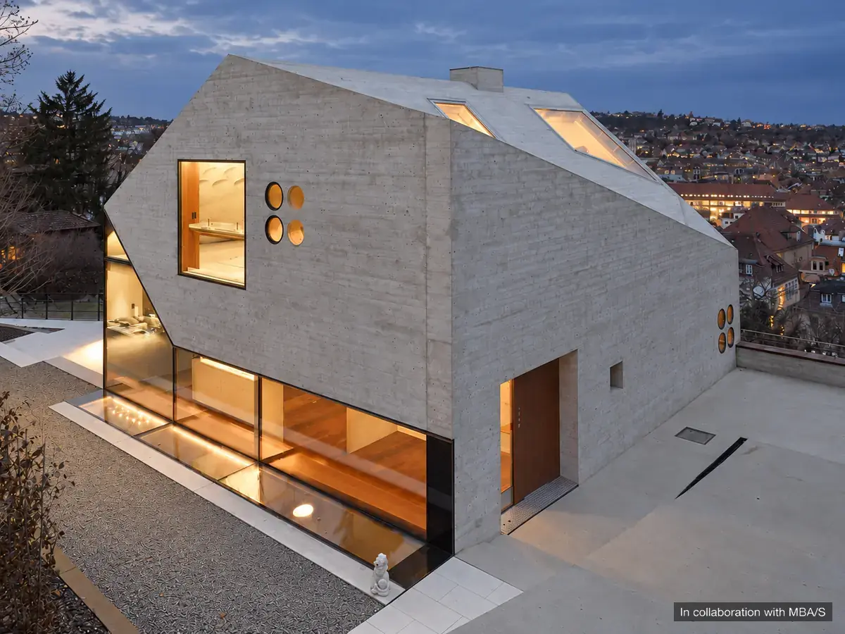 Angular concrete house with broad glazing and warm interior light at dusk. In collaboration with MBA/S.